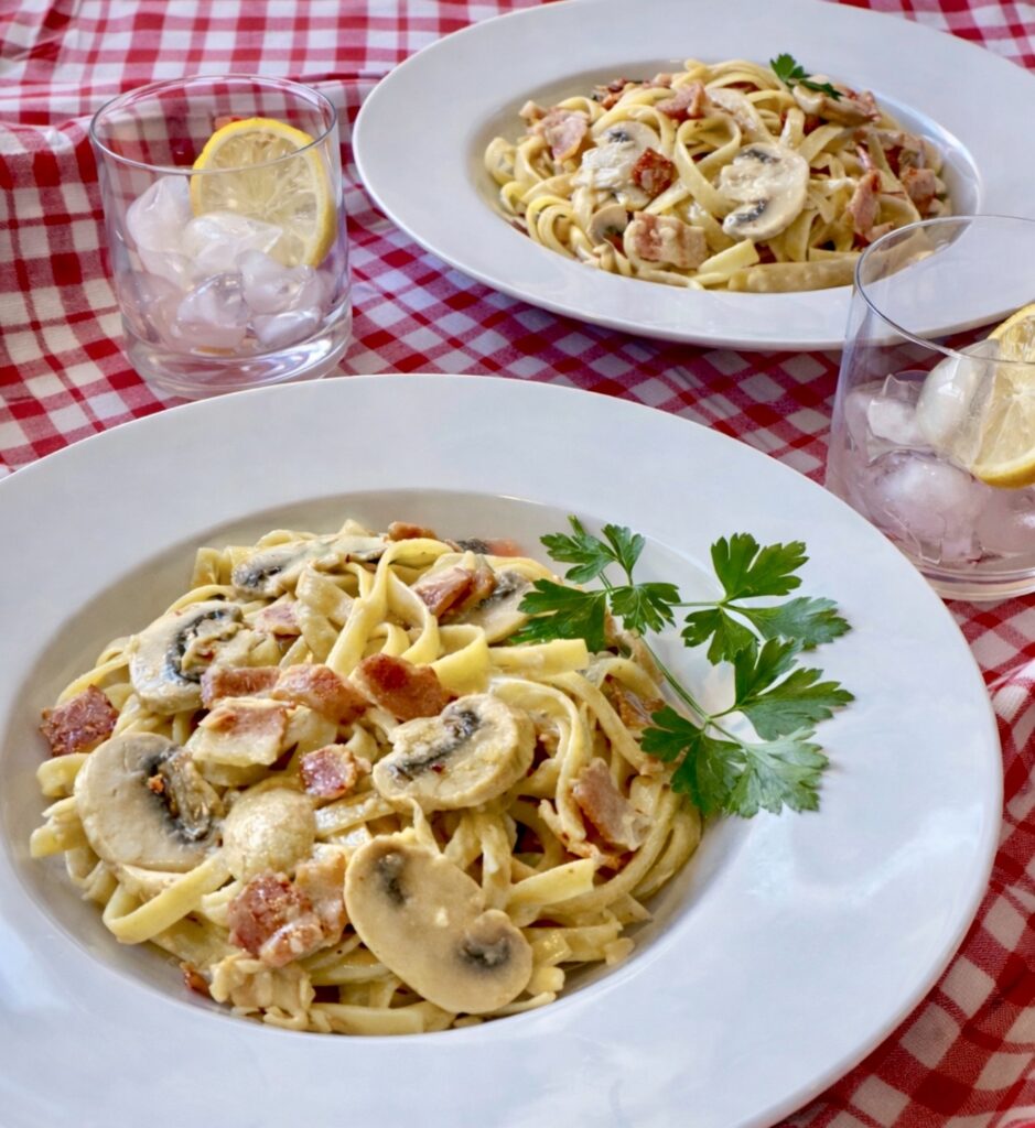 Overhead view of two plates of Fettuccine Boscaiola with creamy mushroom and bacon sauce on a red checkered tablecloth
