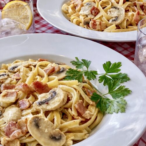 Two bowls of Fettuccine Boscaiola with mushrooms and bacon served on a red gingham tablecloth with iced lemon water