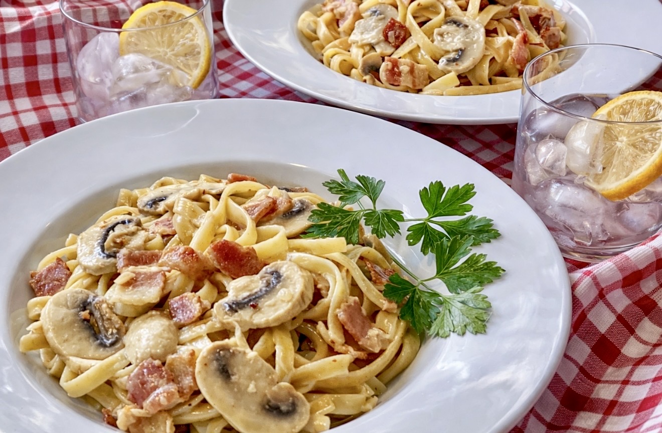 Two bowls of Fettuccine Boscaiola with mushrooms and bacon served on a red gingham tablecloth with iced lemon water