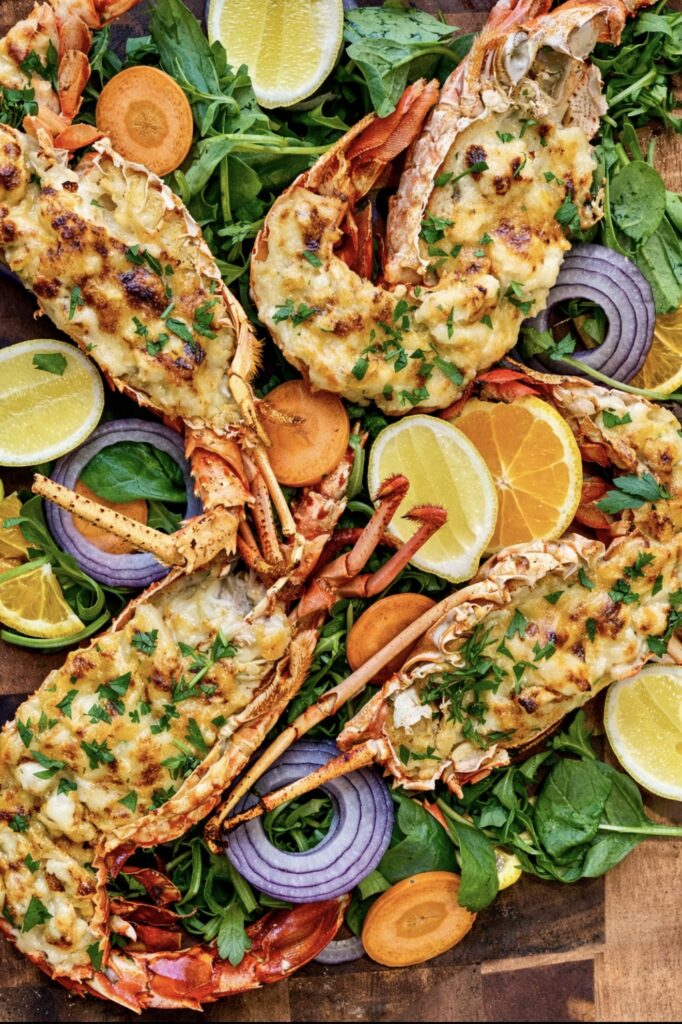 Baked lobster mornay halves topped with creamy cheese sauce, served with greens, citrus slices and red onion on a wooden board