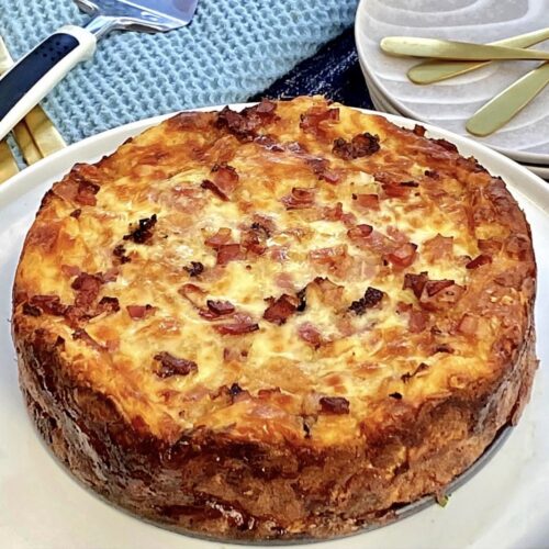 Overnight strata with eggs bacon and cheese baked until golden, served whole on a white plate with a crisp browned crust and melted cheese topping.