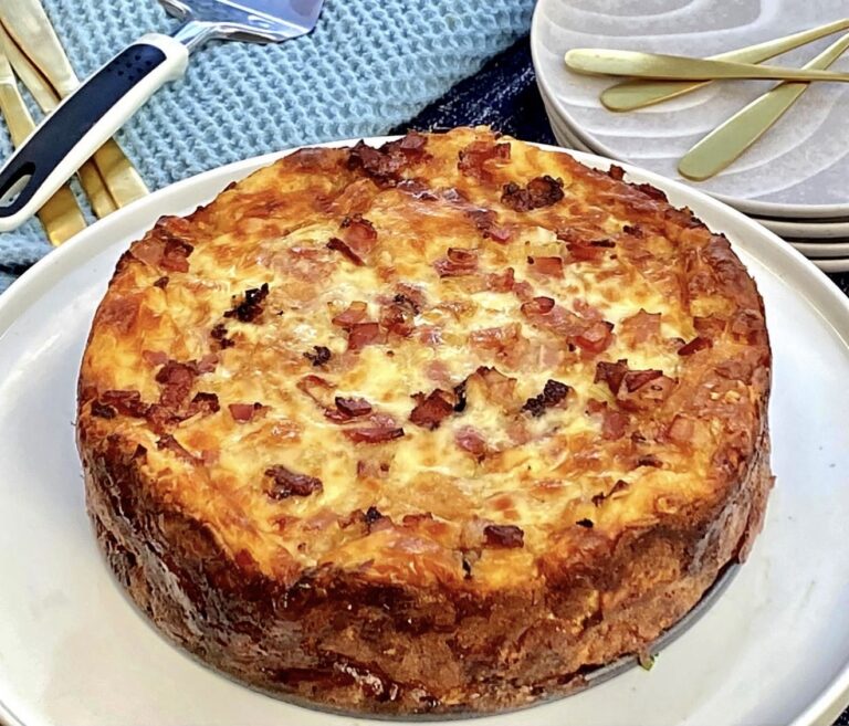 Overnight strata with eggs bacon and cheese baked until golden, served whole on a white plate with a crisp browned crust and melted cheese topping.
