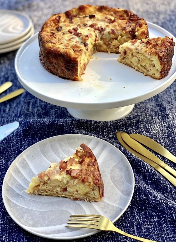Overnight strata with eggs bacon and cheese displayed on a white cake stand, with a single slice served on a plate showing the fluffy layered crum