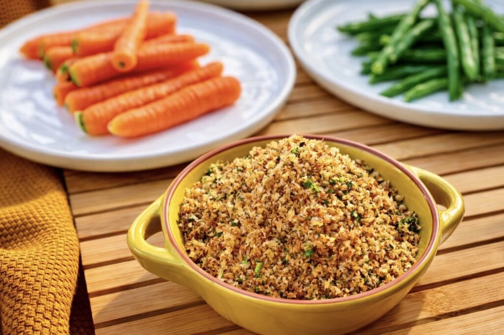 Anchovy breadcrumbs in a yellow ceramic dish served with carrots and green beans on a wooden board