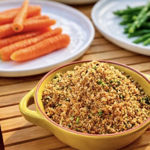 Anchovy breadcrumbs in a yellow ceramic dish served with carrots and green beans on a wooden board