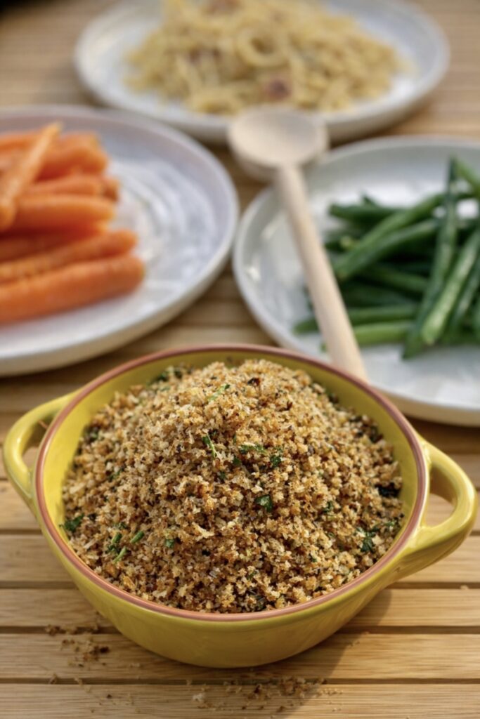 Anchovy breadcrumbs served in a yellow bowl with carrots and green beans in the background