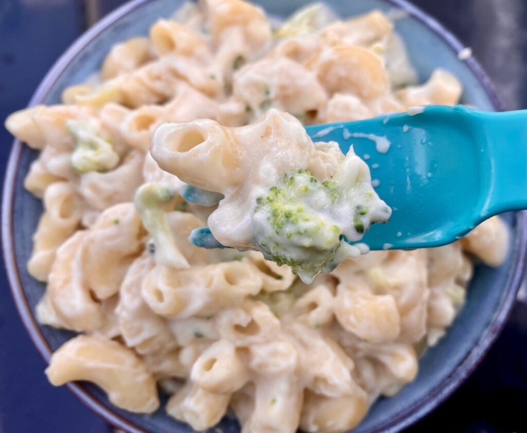 Close-up of baby mac and cheese with broccoli on a blue spoon, showing soft pasta and creamy sauce texture