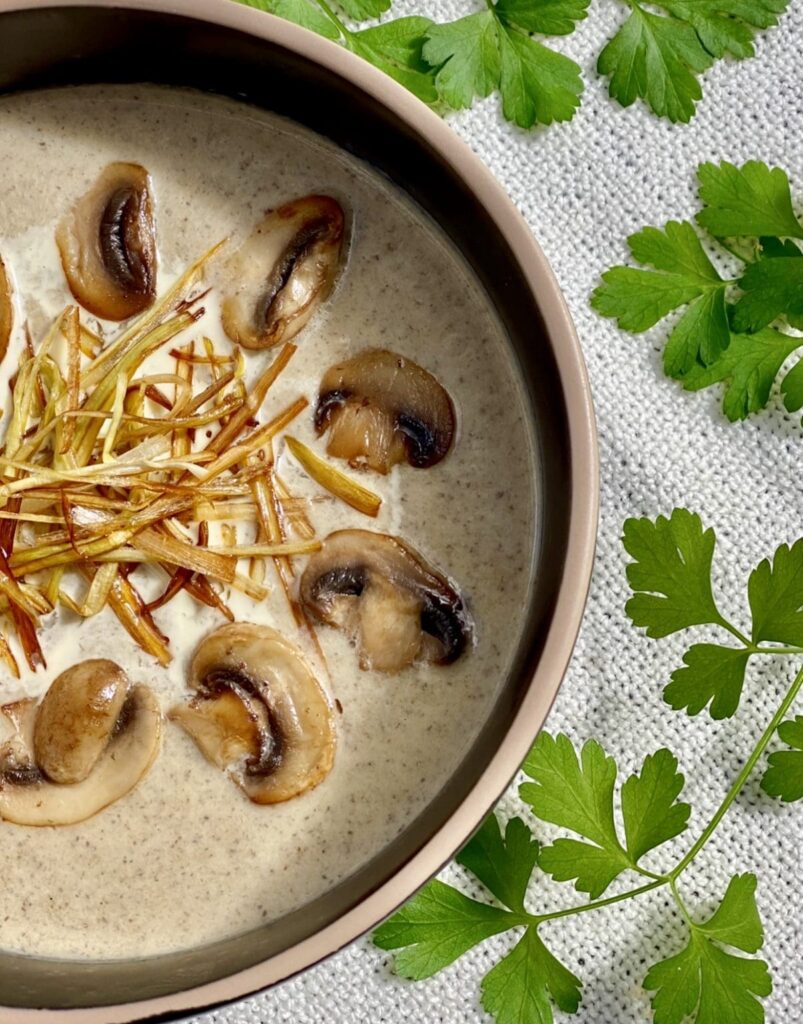 Close up of creamy mushroom leek soup topped with sautéed mushrooms and crispy leeks, served in a ceramic bowl on a light cloth with fresh parsley leaves.