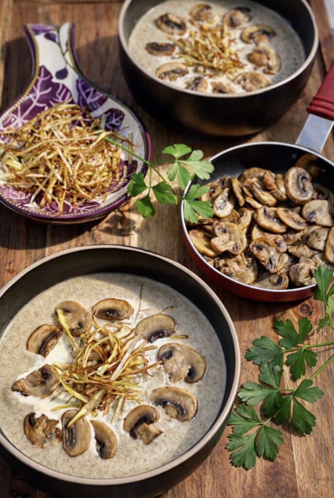 Creamy mushroom leek soup served in bowls, topped with sautéed mushrooms and crispy fried leeks, styled on a wooden board with extra mushrooms and leeks on the side.