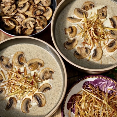 Overhead view of mushroom leek soup in ceramic bowls, garnished with caramelised mushrooms and fried leeks, with a pan of sautéed mushrooms and extra leek garnish alongside.