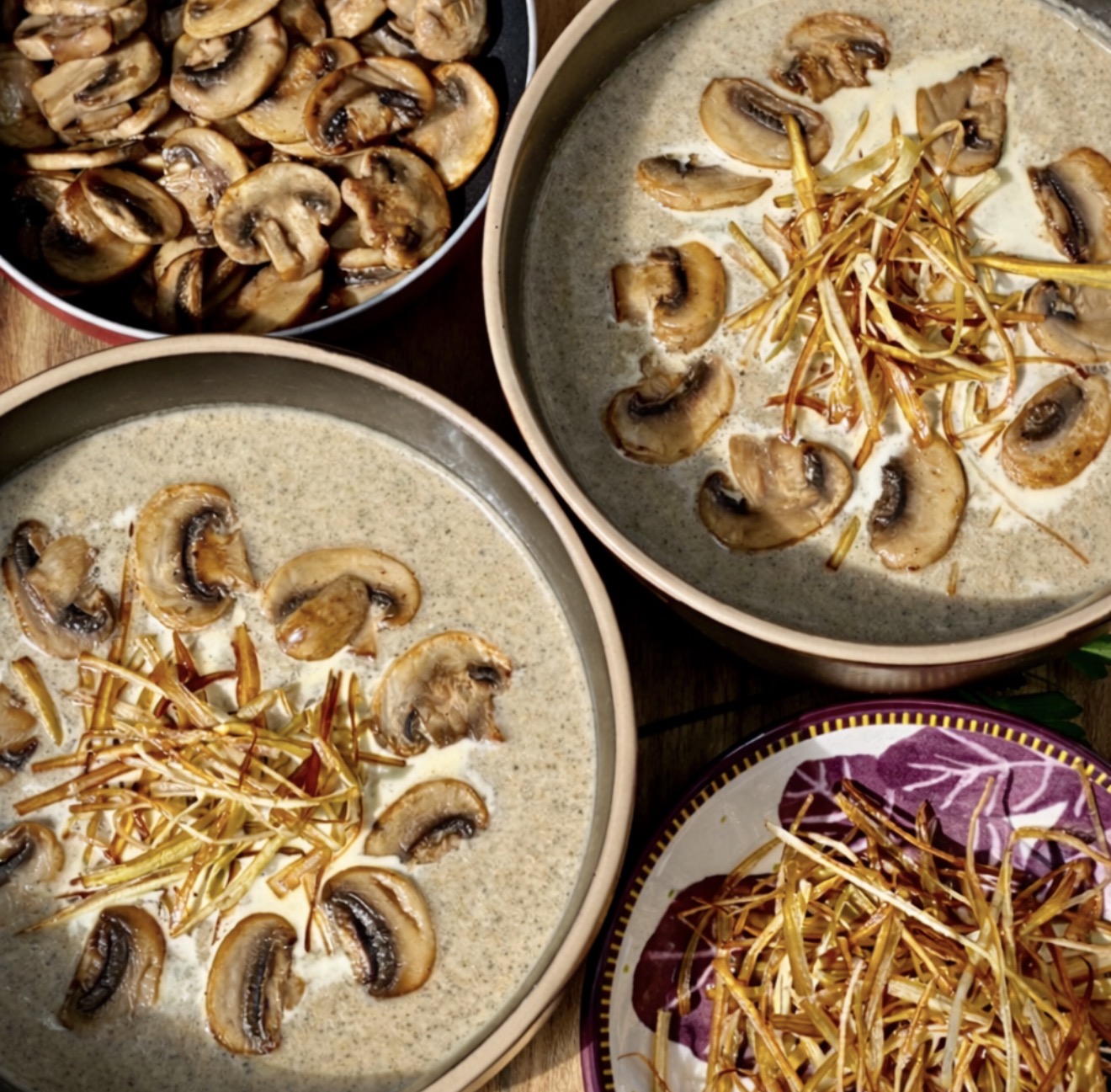 Overhead view of mushroom leek soup in ceramic bowls, garnished with caramelised mushrooms and fried leeks, with a pan of sautéed mushrooms and extra leek garnish alongside.