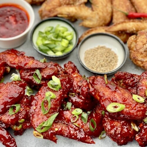 Spicy Korean chicken wings coated in a glossy red gochujang sauce, garnished with sliced spring onions, served with sesame seeds and dipping sauces on a tray.