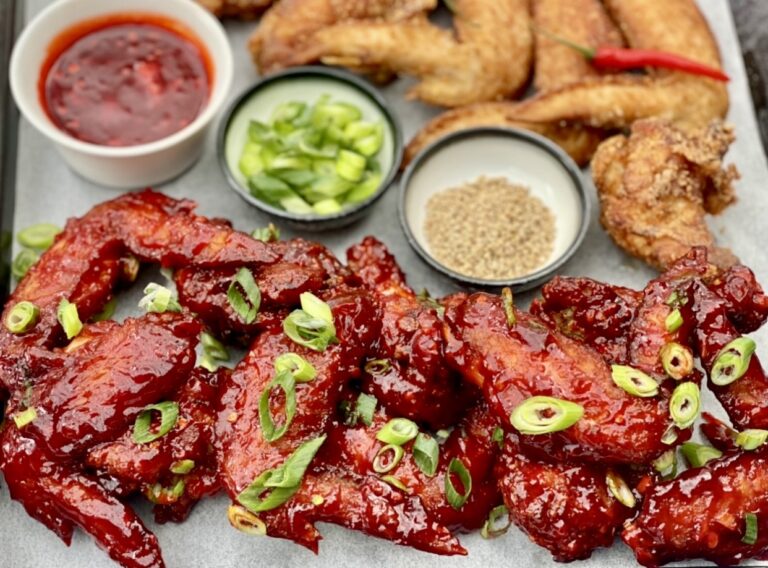 Spicy Korean chicken wings coated in a glossy red gochujang sauce, garnished with sliced spring onions, served with sesame seeds and dipping sauces on a tray.