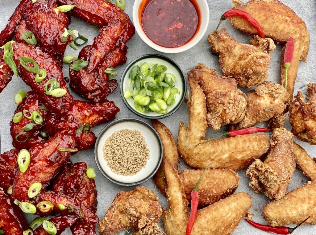 Overhead view of spicy Korean chicken wings and crispy fried wings arranged on baking paper with bowls of gochujang sauce, spring onions, and sesame seeds.