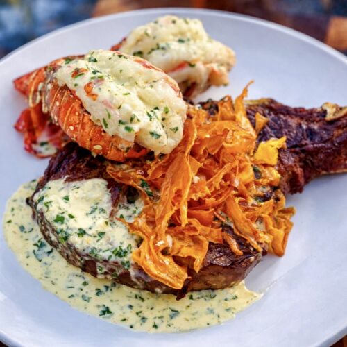 Surf and turf with lobster featuring grilled steak topped with creamy herb sauce, lobster tails, and crispy sweet potato garnish on a white plate