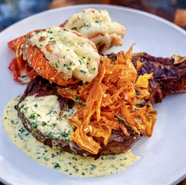 Surf and turf with lobster featuring grilled steak topped with creamy herb sauce, lobster tails, and crispy sweet potato garnish on a white plate