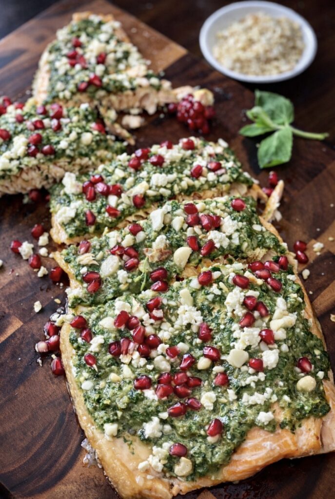 Roasted salmon fillet topped with fresh salsa verde, crumbled feta, macadamia nuts and pomegranate seeds on a wooden board.
