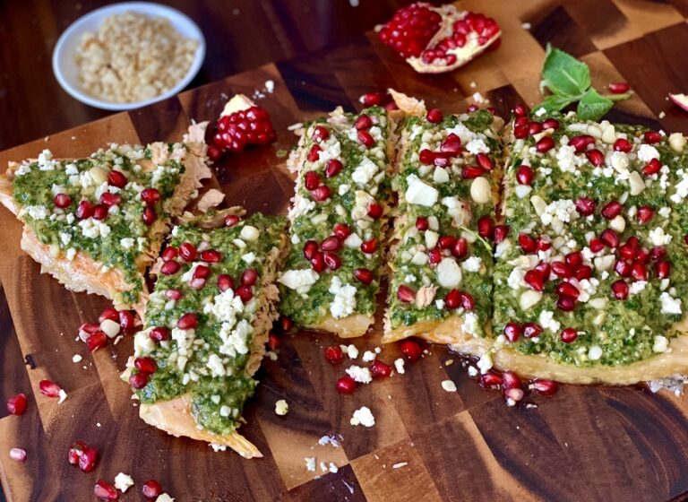 Salmon with salsa verde served on a wooden cutting board, garnished with feta, macadamia nuts and vibrant pomegranate arils.