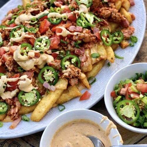 Plate of loaded fries with sausage, bacon and jalapeños served with creamy cheesy sauce and fresh tomato salsa.