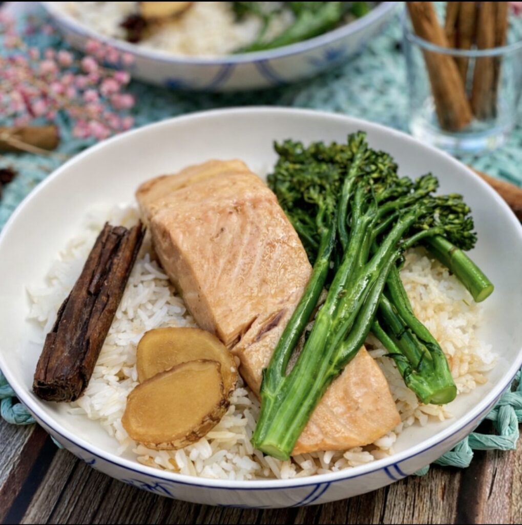 Poached Salmon in Chinese spiced brothserved over fluffy white rice with broccolini, ginger slices and cinnamon, styled in a white bowl.Braised salmon fillet served over fluffy white rice with broccolini, ginger slices and cinnamon, styled in a white bowl.