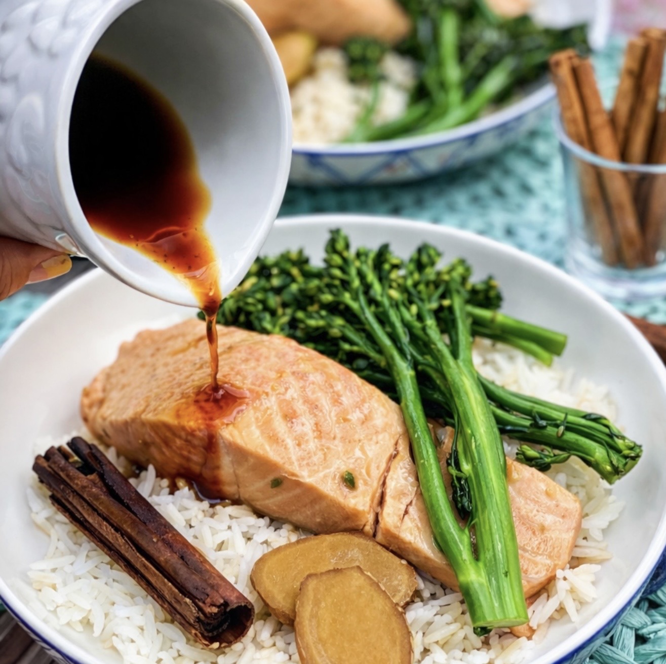 Soy sauce being poured over tender Poached Salmon in Chinese spiced broth with broccolini and jasmine rice, garnished with ginger slices and cinnamon stick.