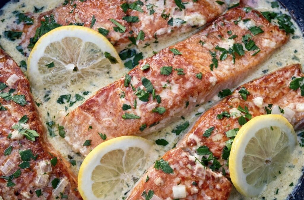 Close up of pan seared salmon in creamy lemon sauce garnished with parsley and sliced lemons.