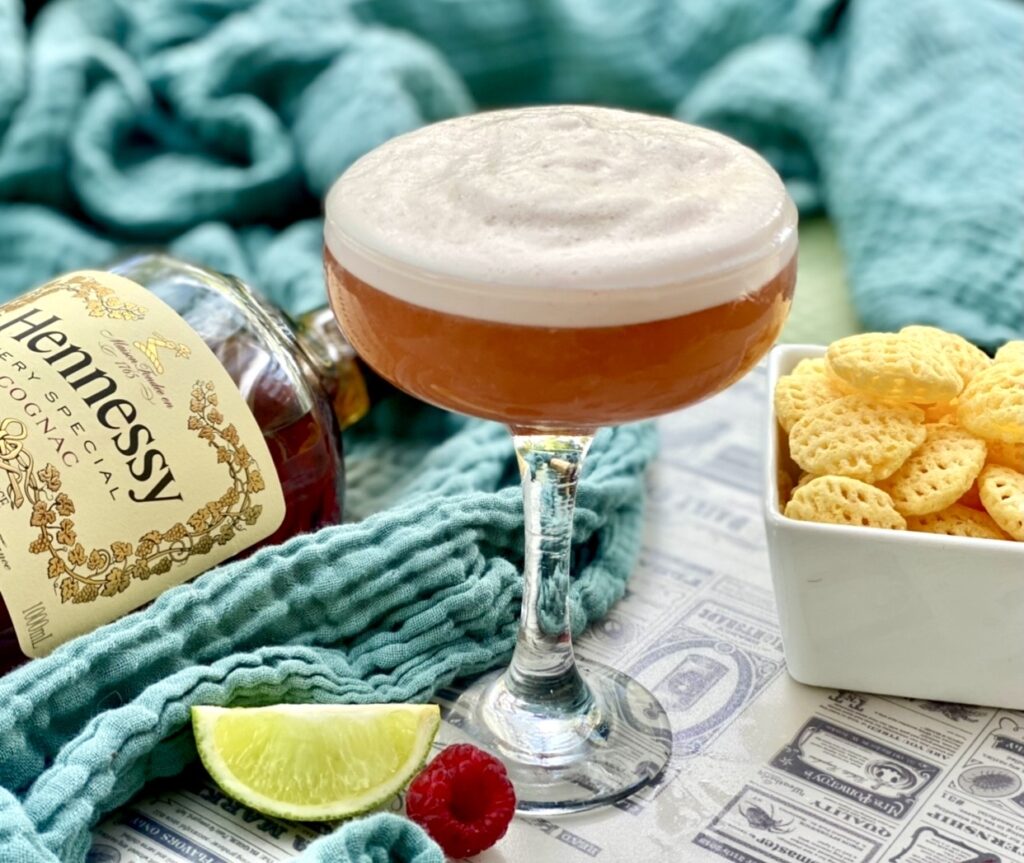 Close up of a frothy pineapple raspberry Hennessy cocktail in a coupe glass with fresh lime and raspberries.
