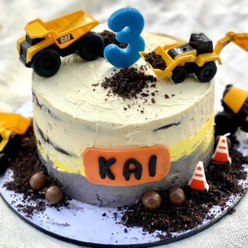 Construction themed birthday cake decorated with yellow toy trucks, chocolate crumb dirt, and a personalised name plaque on the front.