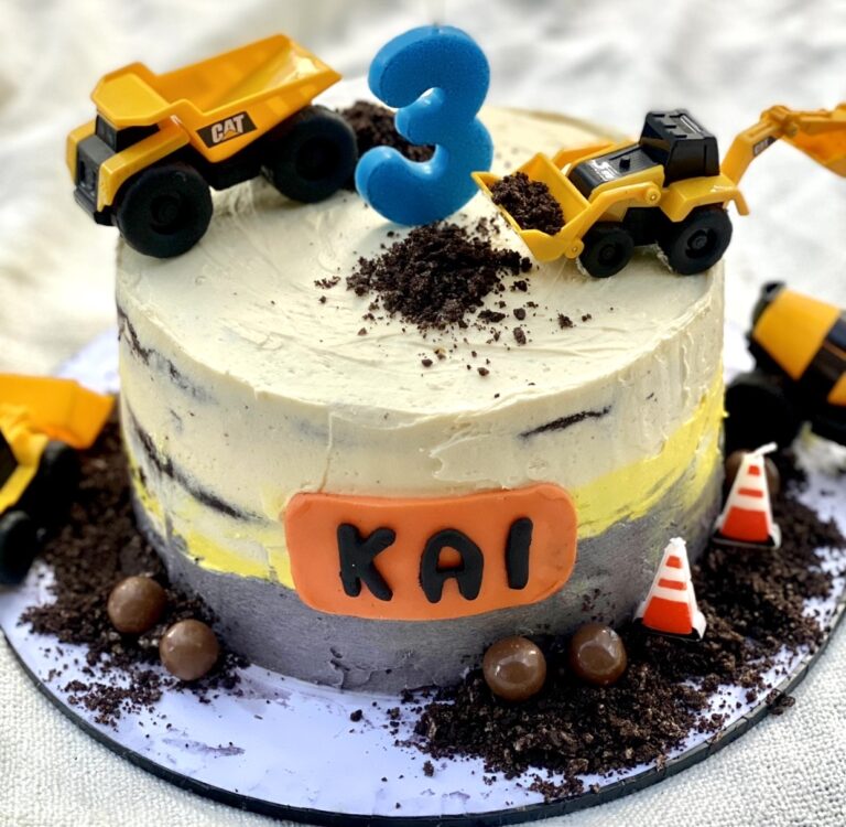 Construction themed birthday cake decorated with yellow toy trucks, chocolate crumb dirt, and a personalised name plaque on the front.