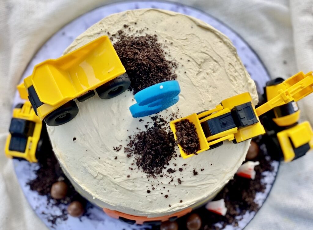 Top view of a construction cake decorated with toy trucks, crushed chocolate cookie dirt, and edible construction details.