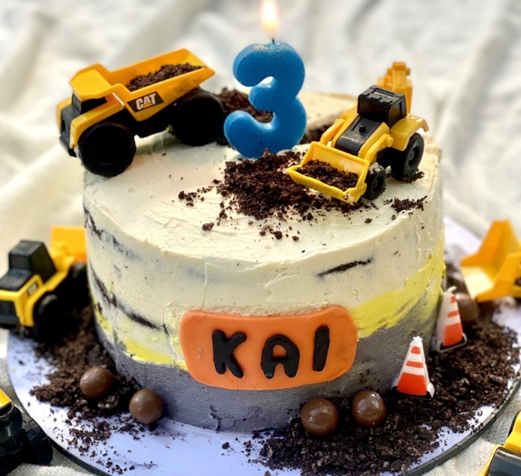 Construction themed cake decorated with toy trucks and personalised name plaque, surrounded by chocolate dirt and construction cones.