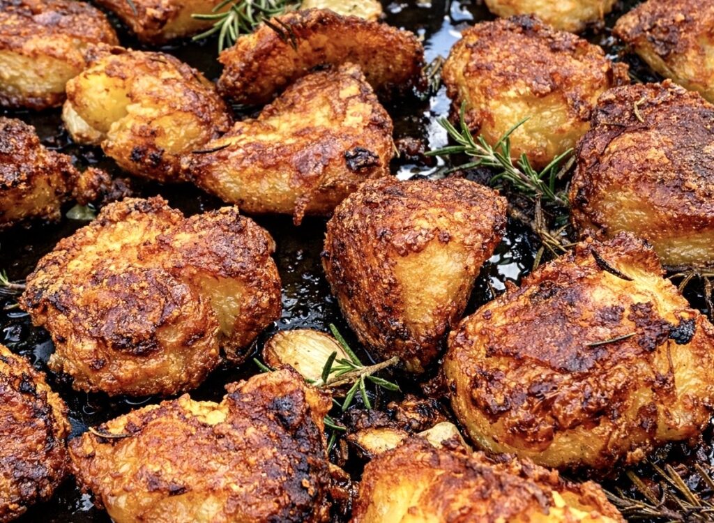 Crispy duck fat potatoes roasting on a black tray with fresh rosemary and garlic cloves, golden and crunchy on the outside.