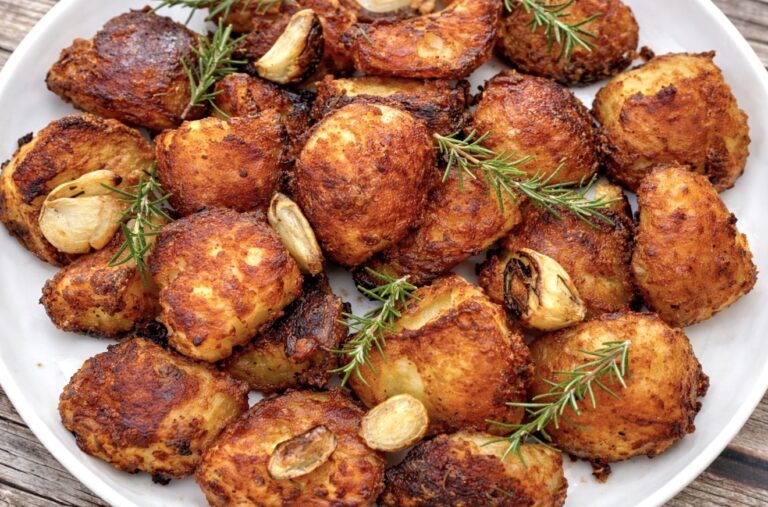 Crispy duck fat potatoes served in a white bowl with roasted garlic and fresh rosemary, perfectly golden and crunchy.