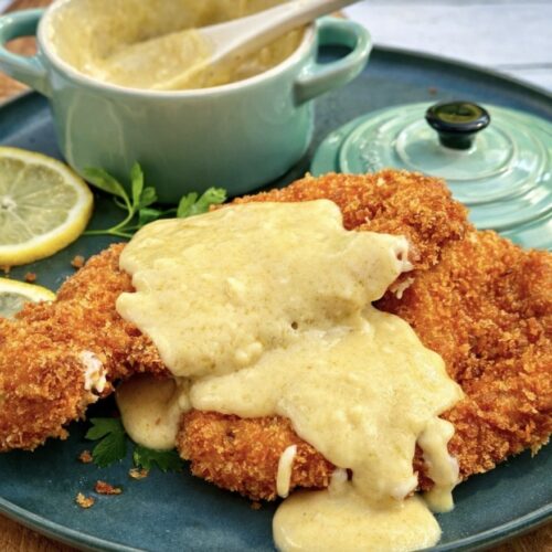 Golden crispy chicken schnitzel topped with creamy cheese sauce, served on a teal plate with lemon slices and fresh parsley.