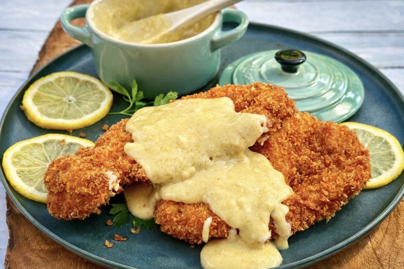 Golden crispy chicken schnitzel topped with creamy cheese sauce, served on a teal plate with lemon slices and fresh parsley.