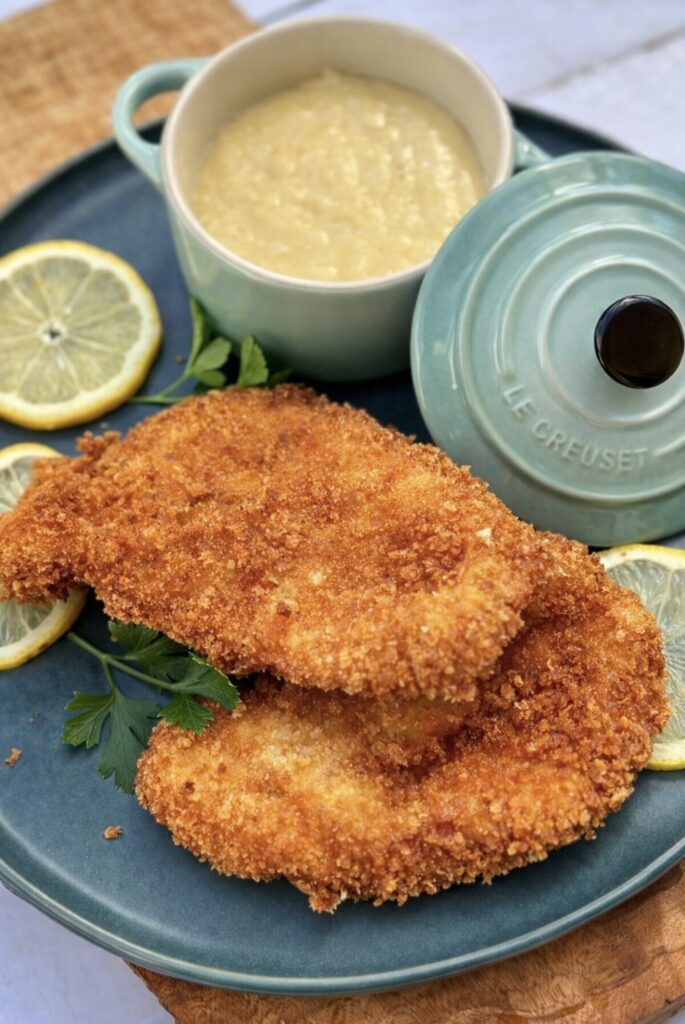 Crispy breaded chicken schnitzel served with a small pot of creamy cheese sauce, lemon slices and parsley on a teal plate.