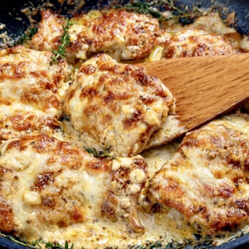 Cheesy chicken thighs simmering in a creamy sauce, with melted golden cheese and herbs, lifted from the pan with a wooden spoon