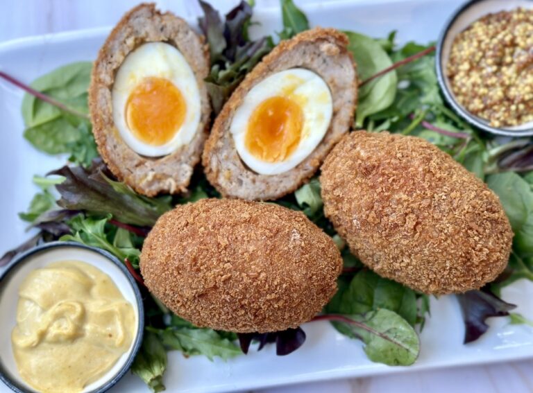 Scotch eggs cut in half showing soft set yolks, coated in golden breadcrumbs and served on a plate with leafy greens and mustard sauce