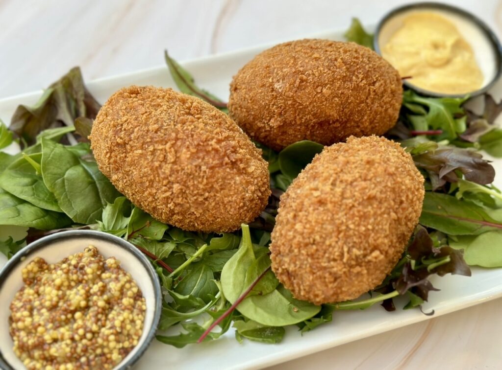 Whole Scotch eggs with crisp golden crumb, served on a platter with fresh greens and a bowl of grainy mustard