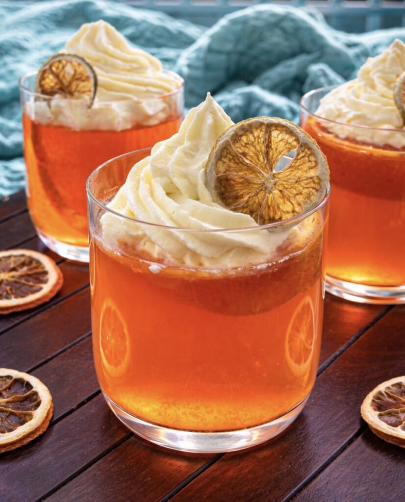 Aperol spritz jelly served in clear glasses, vibrant orange jelly topped with whipped cream and dried citrus slices on a wooden table.