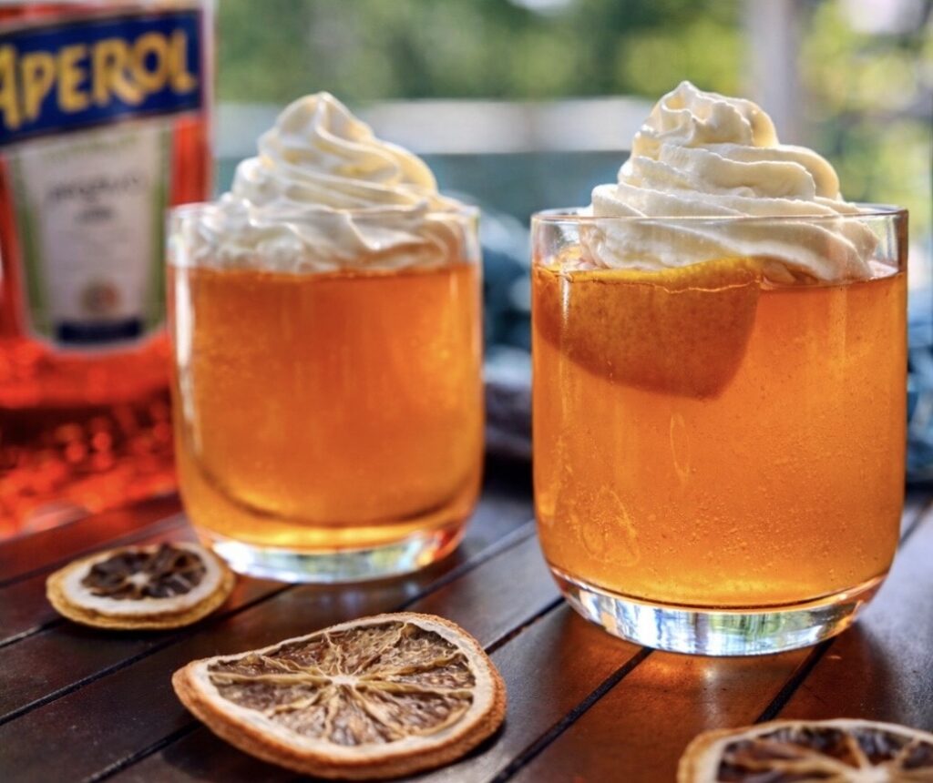 Aperol spritz jelly topped with whipped cream in clear glasses, styled with dried citrus slices and aperol bottles on a wooden table.