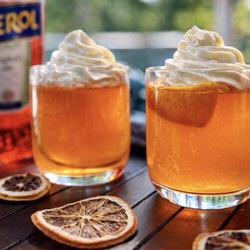 Aperol spritz jelly topped with whipped cream in clear glasses, styled with dried citrus slices and aperol bottles on a wooden table.