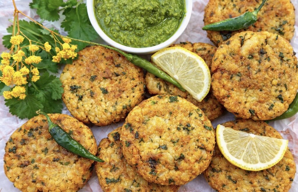 Golden vegan aloo tikki Indian potato patties served with green coriander chutney, fresh green chillies, lemon wedges, and herbs on parchment paper.