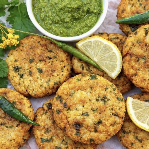 Golden vegan aloo tikki Indian potato patties served with green coriander chutney, fresh green chillies, lemon wedges, and herbs on parchment paper.