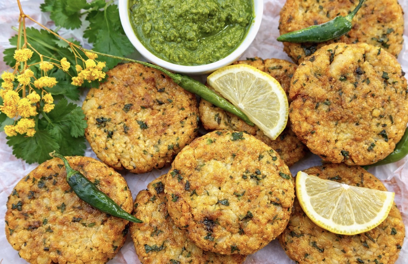 Golden vegan aloo tikki Indian potato patties served with green coriander chutney, fresh green chillies, lemon wedges, and herbs on parchment paper.