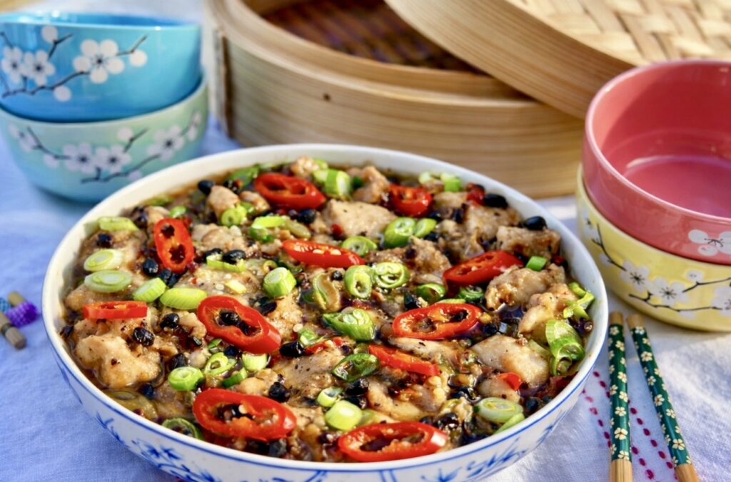 Chinese steamed black bean chicken served in a shallow bowl, topped with sliced red chilli, spring onions and fermented black beans, with bamboo steamers in the background