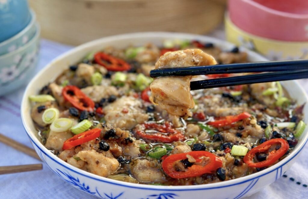 Close up of Chinese steamed black bean chicken with a piece of tender chicken lifted by chopsticks, showing the rich black bean sauce, red chilli and spring onions