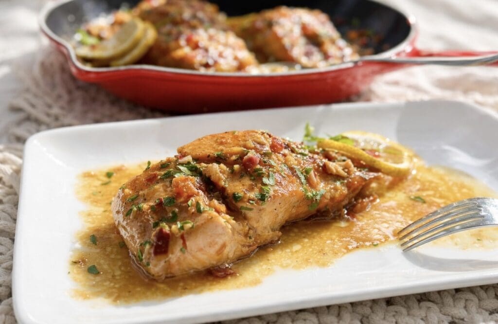 A fillet of Honey Garlic Butter Salmon on a rectangle plate covered in mouthwatering sauce with a fork and pan in the background.