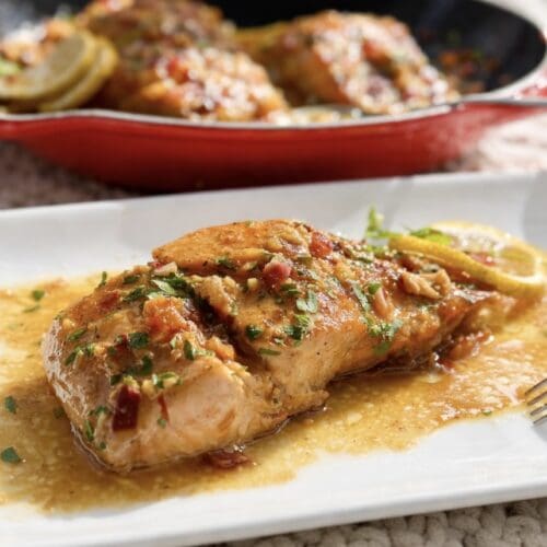 A fillet of Honey Garlic Butter Salmon on a rectangle plate covered in mouthwatering sauce with a fork and pan in the background.