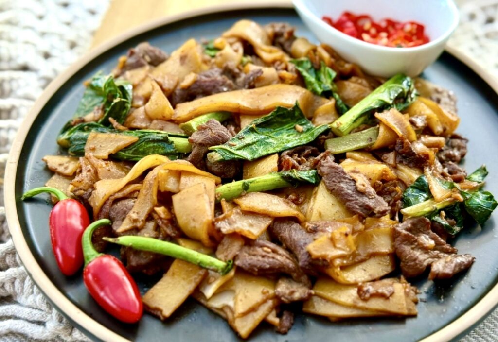 Pad see ew with wide rice noodles, tender beef, Chinese broccoli and a glossy soy based sauce, served on a dark plate